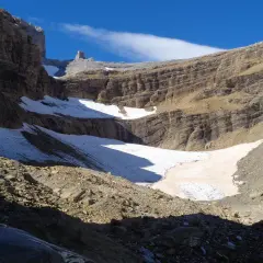 Un paisaje montañoso con nieve y rocas, donde el sol ilumina la **col de tentes** y el cielo azul.