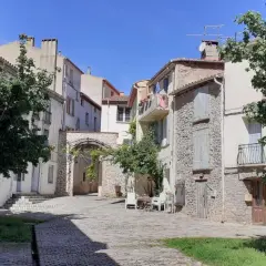 Calle adoquinada en Arles-sur-Tech, con casas de piedra y fachadas blancas, un arco de piedra y árboles frondosos bajo un cielo azul.