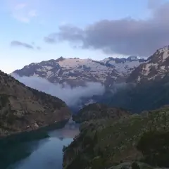 Paisaje montañoso con un lago sereno y picos nevados. Nubes bajas envuelven las montañas, creando una atmósfera mística. Un lugar idílico para explorar, como el que se encuentra en Besiberri Sud.