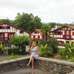 Mujer sentada en un muro de piedra, con casas blancas y rojas típicas del Pirineo Vasco al fondo.