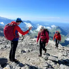 Tres excursionistas suben por una cresta rocosa en el **alt Pirineu**. El sol brilla en un cielo azul claro, con vistas a un vasto paisaje montañoso.