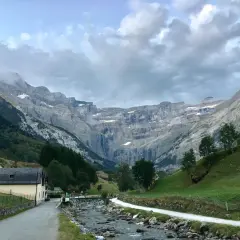 Un camino serpentea junto a un río rocoso hacia el imponente pico Taillón, con una cascada visible en la distancia y un pueblo a la izquierda.