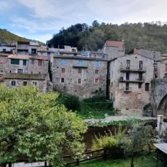 Casas de piedra y un puente medieval en **la Cellera de Ter**, rodeadas de vegetación y colinas boscosas bajo un cielo nublado.