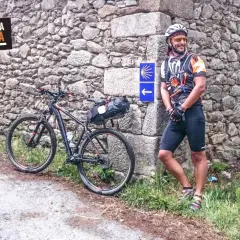 Ciclista sonriente junto a su bici en un camino primitivo, con señal de flecha y concha de peregrino.