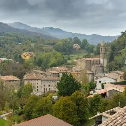 Vista panorámica de Vallfogona del Ripollès, un pueblo rodeado de bosques otoñales y montañas neblinosas. Destaca la iglesia con su campanario.