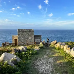 Antigua estaca de bares de piedra junto al mar, con un camino de grava y un fotógrafo capturando la vista.