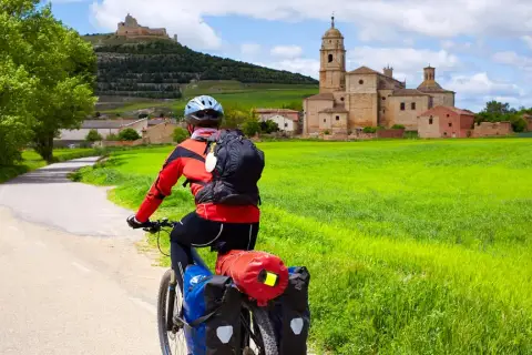 Ciclista en el Camino de Santiago en bici, con un pueblo y un castillo al fondo, rodeado de campos verdes.