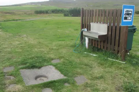 Zona de vaciado de aguas negras para caravana. Un lavabo y un cubo de basura junto a una valla de madera.