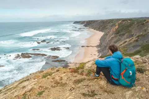Persona con mochila azul contempla la costa salvaje durante un senderismo Portugal. Olas rompen en la playa desierta.