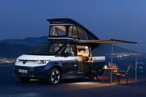Autocaravana Volkswagen California lista para la aventura nocturna, con techo elevable desplegado y toldo extendido sobre un paisaje de luces urbanas.