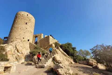 Personas suben escaleras hacia el histórico castillo de Burriac, una torre de piedra antigua bajo un cielo azul claro.