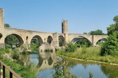 Puente medieval de Besalú, parte de una ruta histórica. Reflejo en el río y vegetación exuberante.