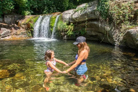 Madre e hija se toman de las manos en la alberca natural, con una cascada al fondo.