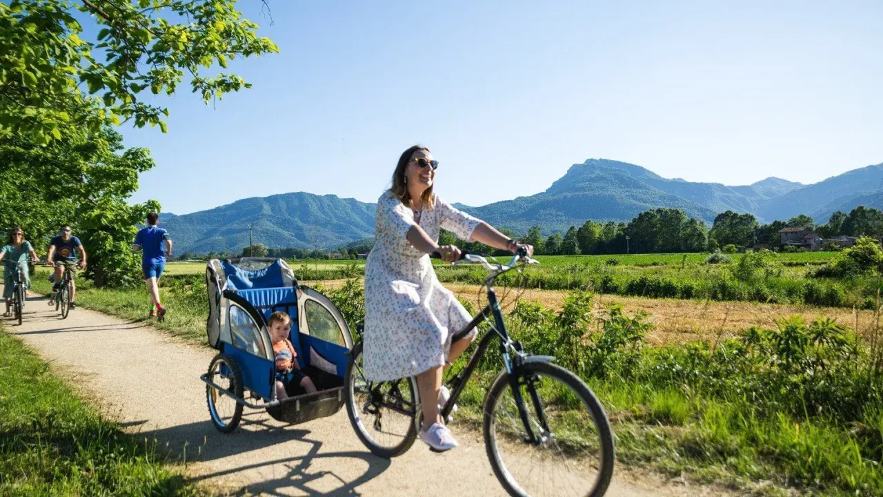 Disfrutando de un día soleado en la ruta el carrilet. Una mujer pedalea con un niño en un remolque, mientras otros ciclistas y un corredor disfrutan del paisaje montañoso.