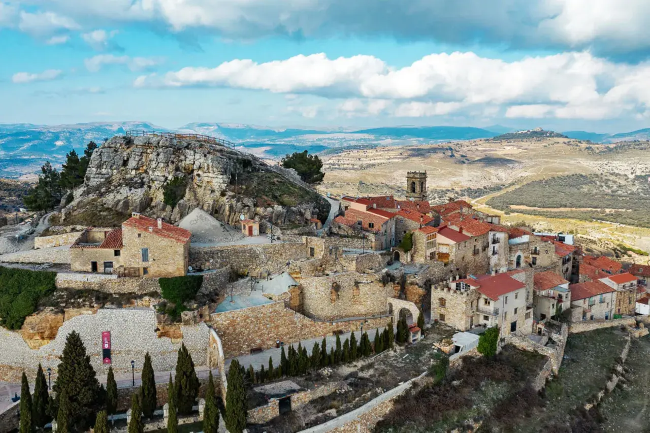 Vista aérea de un pueblo de casas de piedra con tejados rojos, anidado en la ladera de la roca del corb, bajo un cielo azul con nubes.