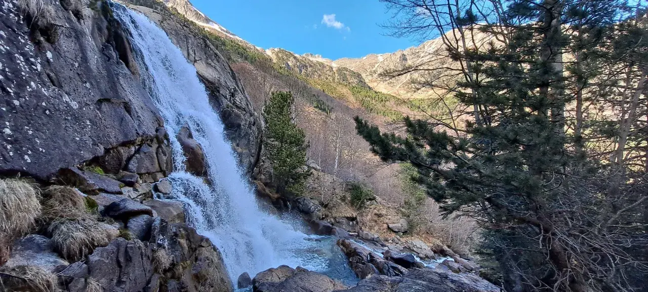 Cascada imponente en un paisaje montañoso, ideal para explorar rutas vielha. El agua cae sobre rocas y vegetación, con árboles y picos nevados al fondo.