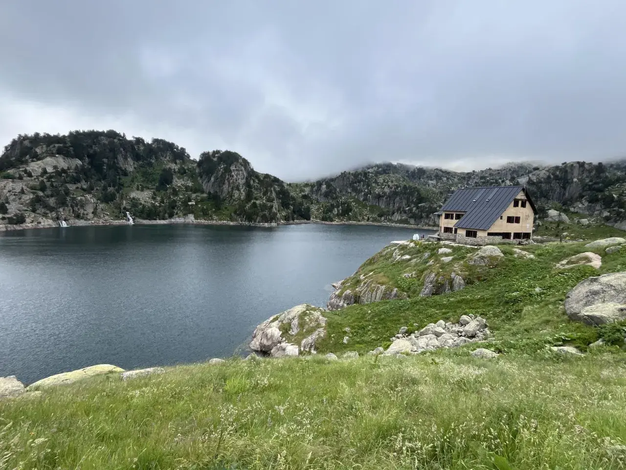 Refugio de montaña junto a un lago rodeado de montañas rocosas y vegetación.