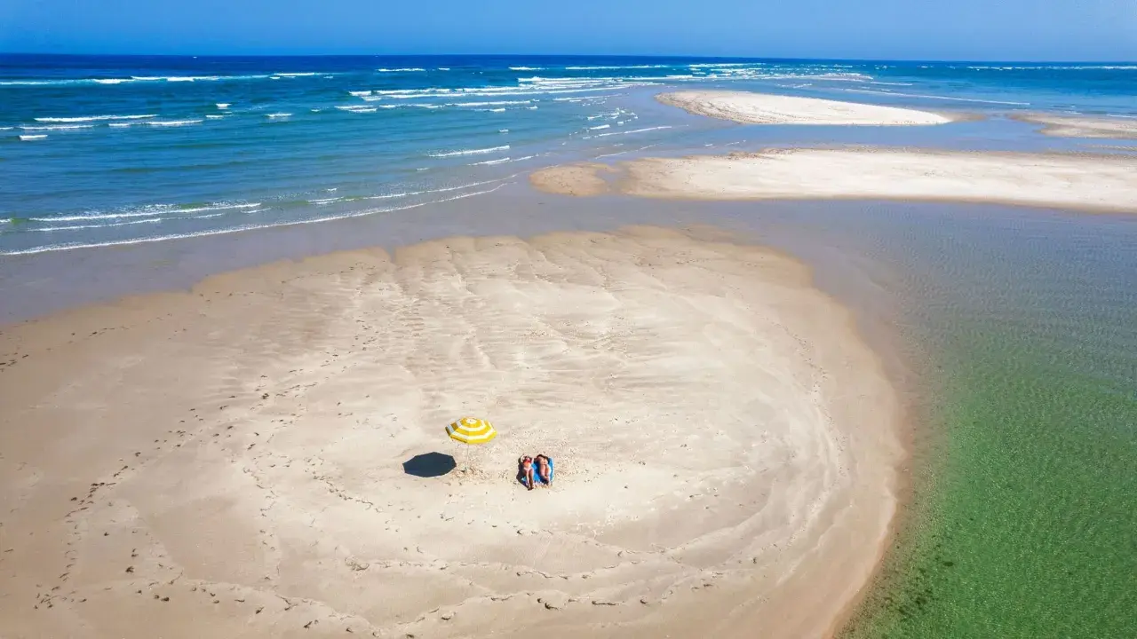 Dos personas descansan bajo una sombrilla a rayas en una playa de arena en Ria Formosa. El mar azul y las aguas cristalinas rodean la isla.