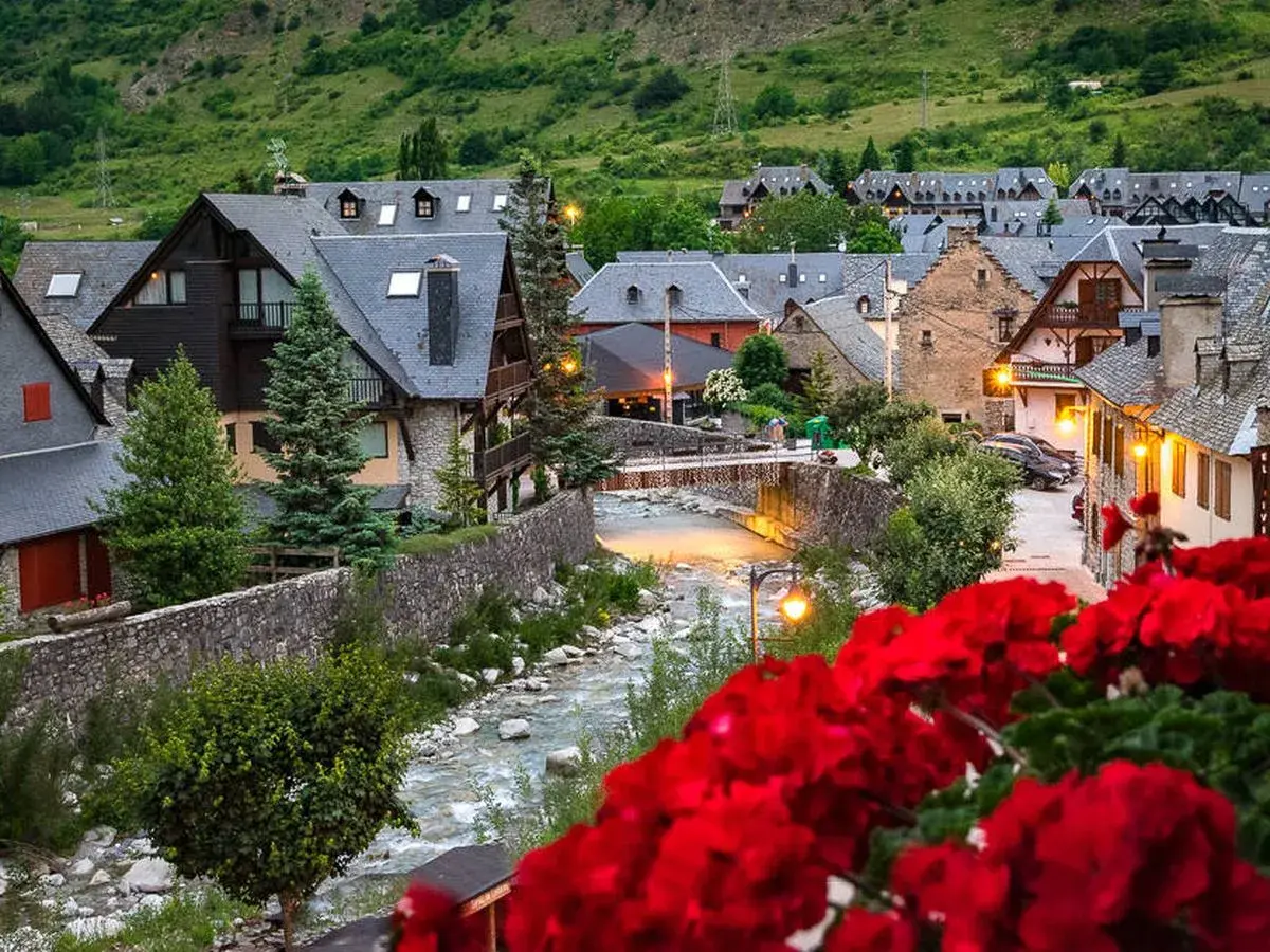 Pueblo pintoresco en el Pirineo Catalán, con casas de piedra y tejados grises junto a un río. Flores rojas en primer plano.