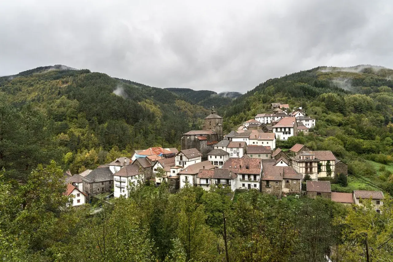 Pueblo pintoresco en el Pirineo Navarro, con casas de piedra y tejados rojos, rodeado de bosques frondosos y montañas nubladas.