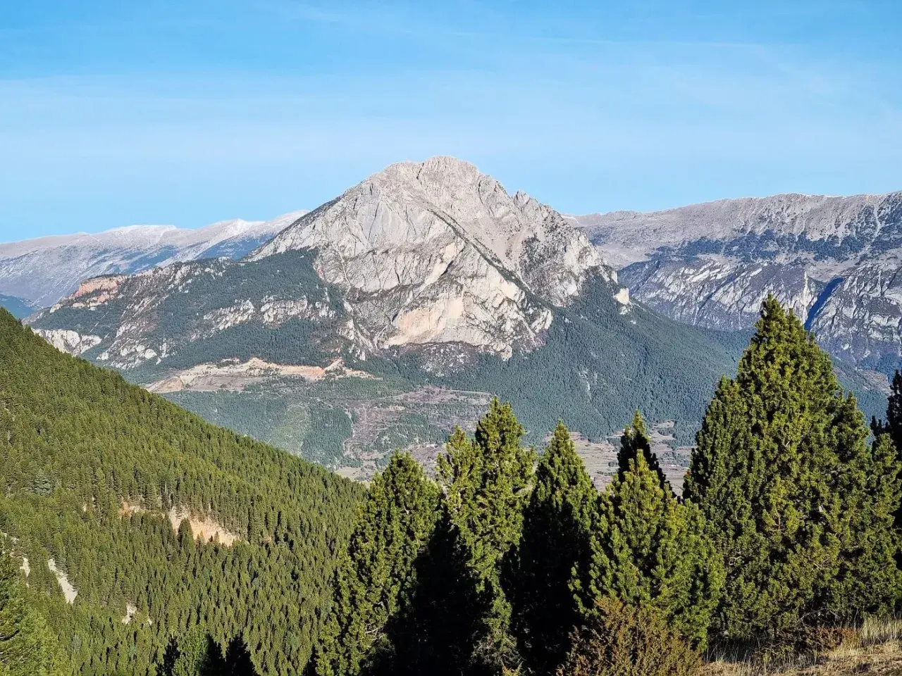 Majestuosa Serra d'Ensija bajo un cielo azul. Bosques de pinos cubren las laderas, con picos rocosos y nevados al fondo.
