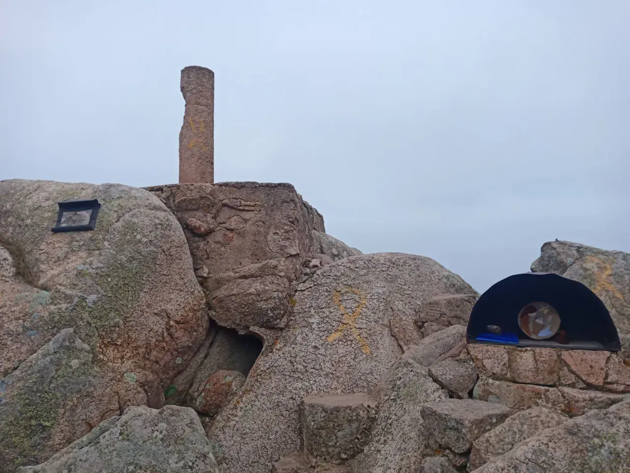 Cima del Puig de Cadiretes con un monolito, rocas y un pequeño refugio con un objeto en su interior.