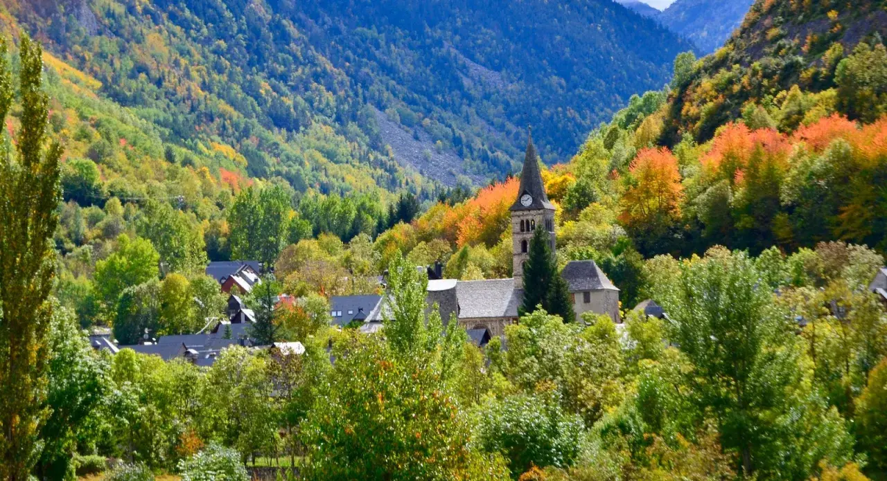 Pueblo en el **Valle de Arán** rodeado de árboles con hojas de otoño en tonos verdes, amarillos y naranjas. Destaca una iglesia con campanario.