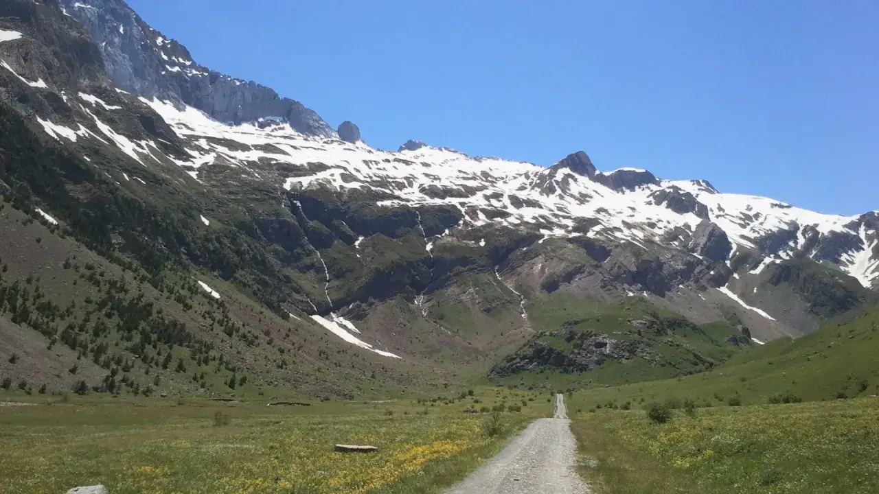 Camino de grava serpentea por el verde valle de Otal, flanqueado por imponentes montañas nevadas bajo un cielo azul claro.