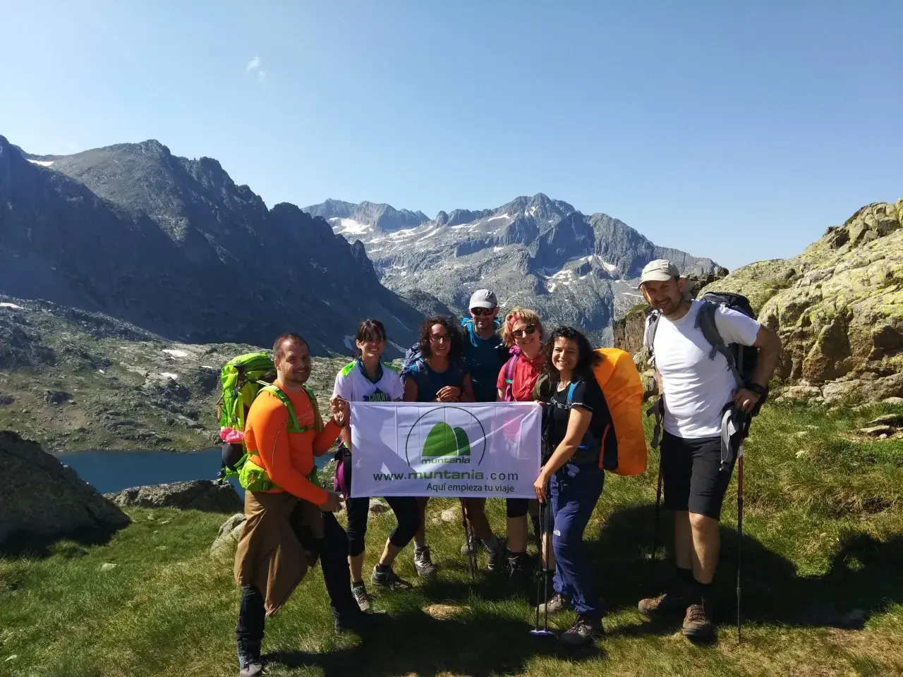 Grupo de excursionistas sonríe en la montaña, listos para la ruta. Llevan mochilas y un cartel que dice "Aquí empieza tu viaje".