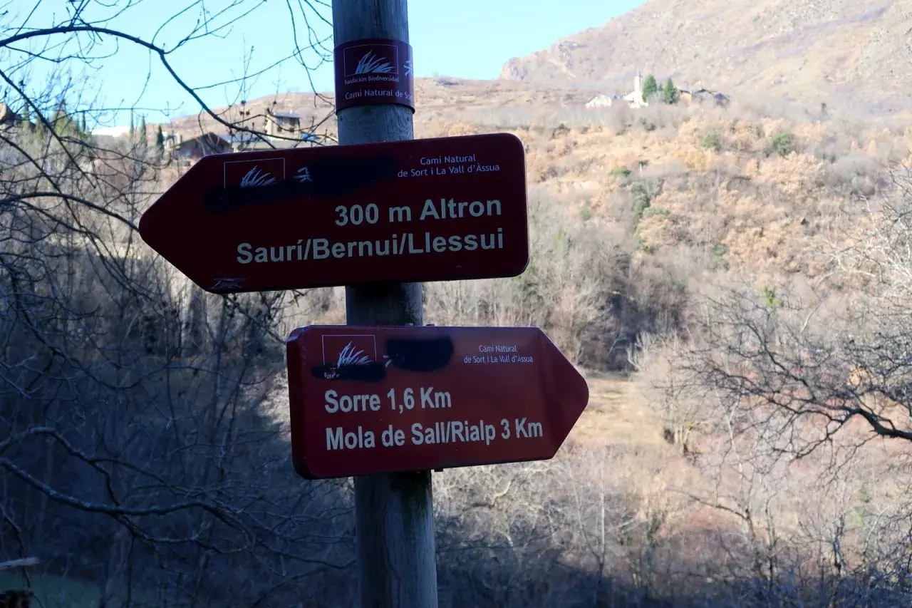 Señales de senderismo en la Vall d'Assua: 300m a Altron y 1,6km a Sorre. El paisaje montañoso y boscoso sugiere una ruta de naturaleza.