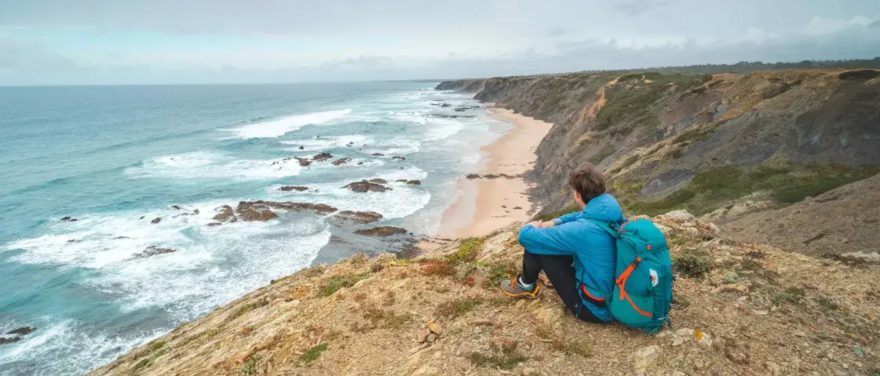 Persona con mochila azul contempla la costa salvaje durante un senderismo Portugal. Olas rompen en la playa desierta.