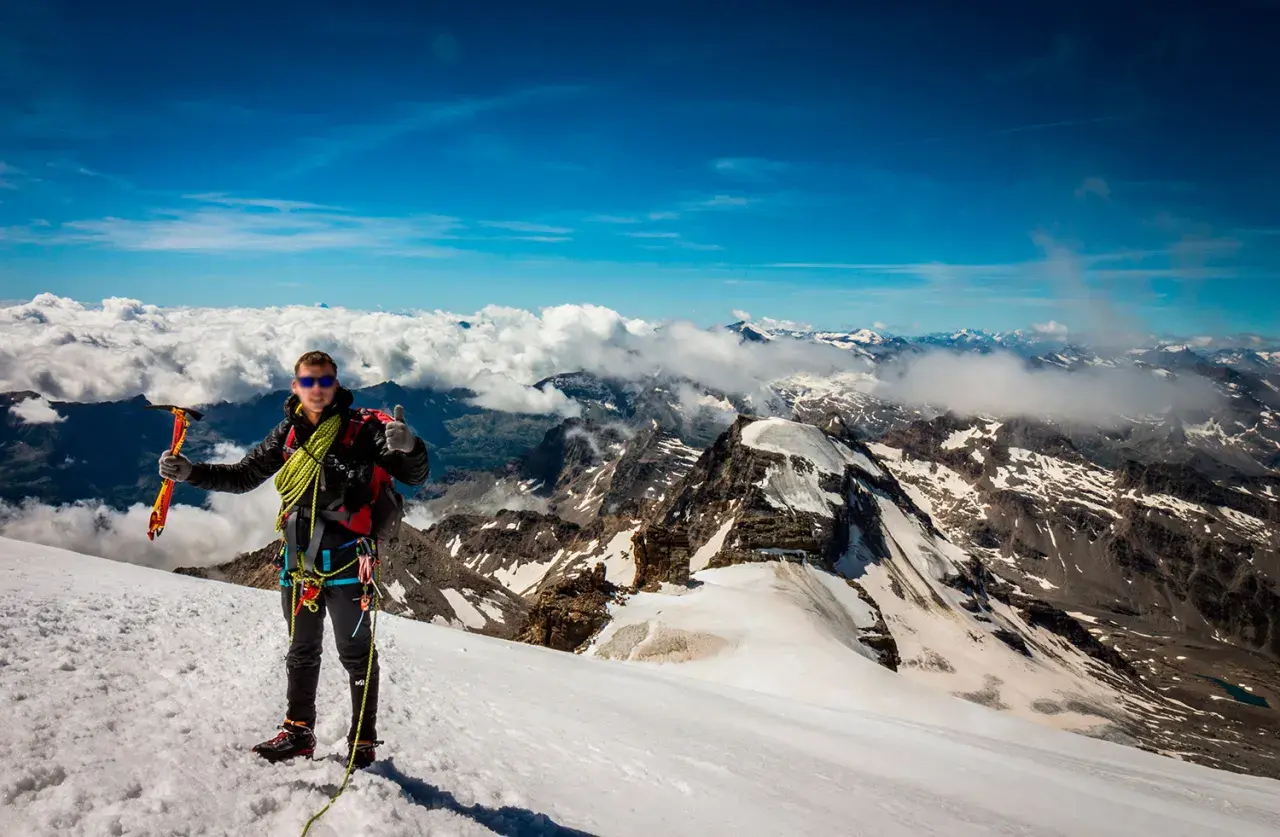 Alpinista en la cima del Gran Paradiso, con piolet y equipo, disfrutando de vistas espectaculares de montañas nevadas y nubes.