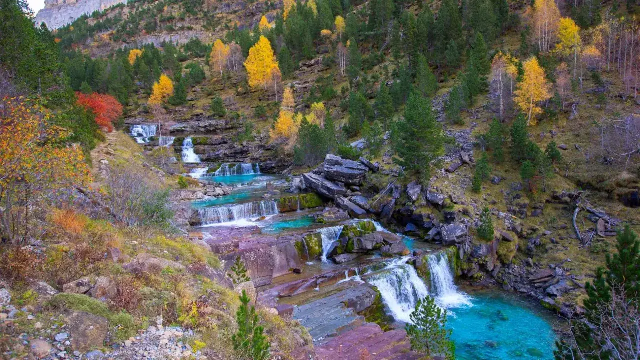 Cascada en las gradas de soaso, con aguas turquesas y rocas rojizas, rodeada de árboles otoñales y pinos verdes.