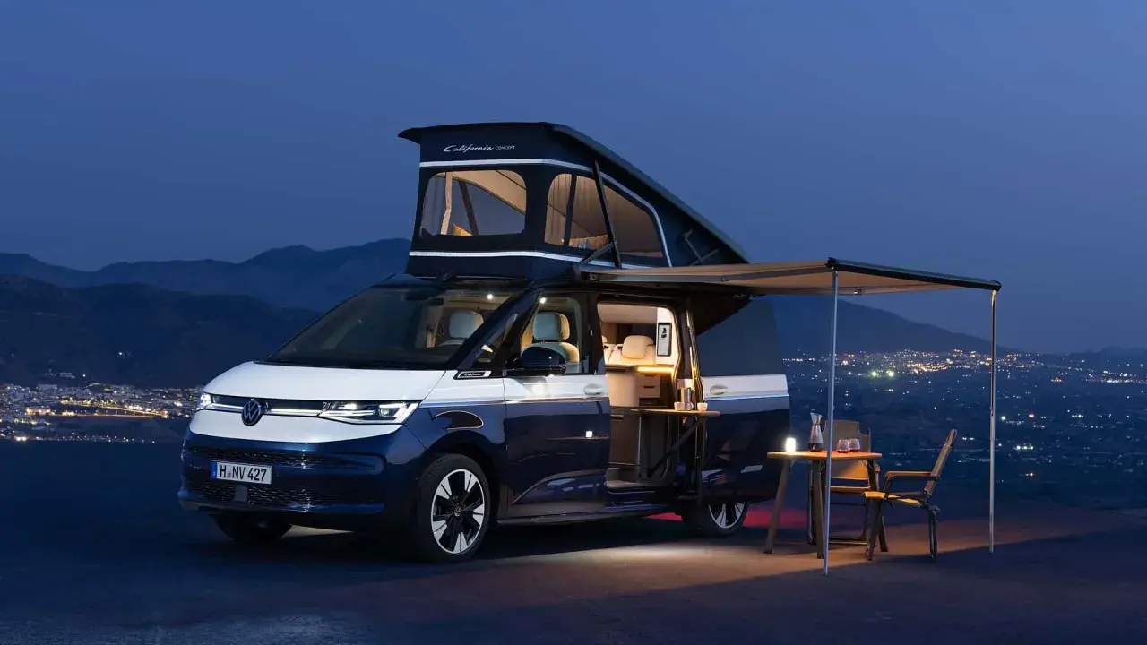 Autocaravana Volkswagen California lista para la aventura nocturna, con techo elevable desplegado y toldo extendido sobre un paisaje de luces urbanas.