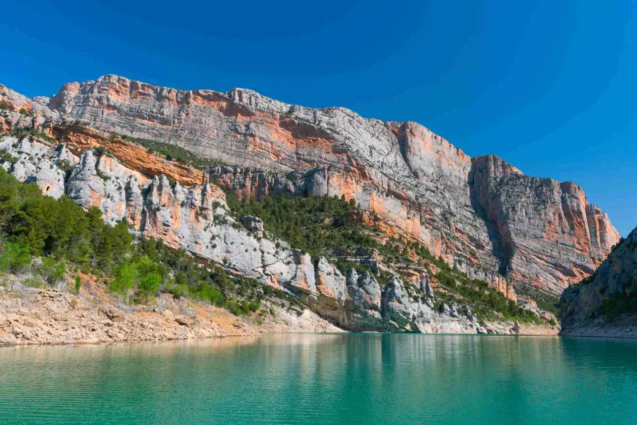Majestuosas paredes rocosas de la sierra del Montsec se reflejan en las aguas turquesas de un embalse.