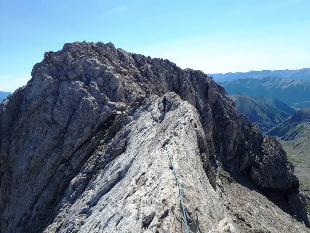 Alpinista en la cresta rocosa del Pico de Vallibierna, con un arnés y cuerda azul. Paisaje montañoso al fondo.