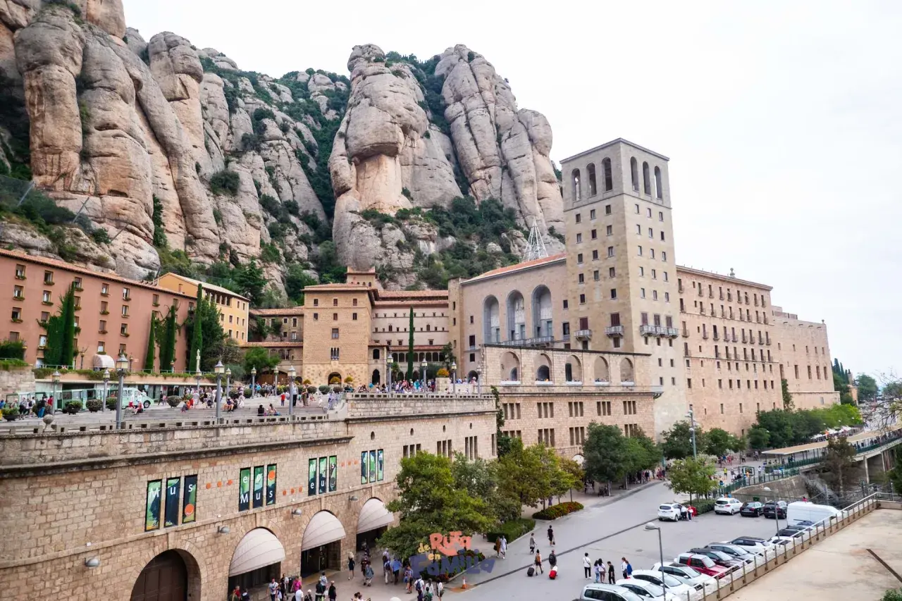 Edificios del monasterio se alzan bajo las imponentes rocas del parc natural de la muntanya de Montserrat. Turistas pasean por la plaza.