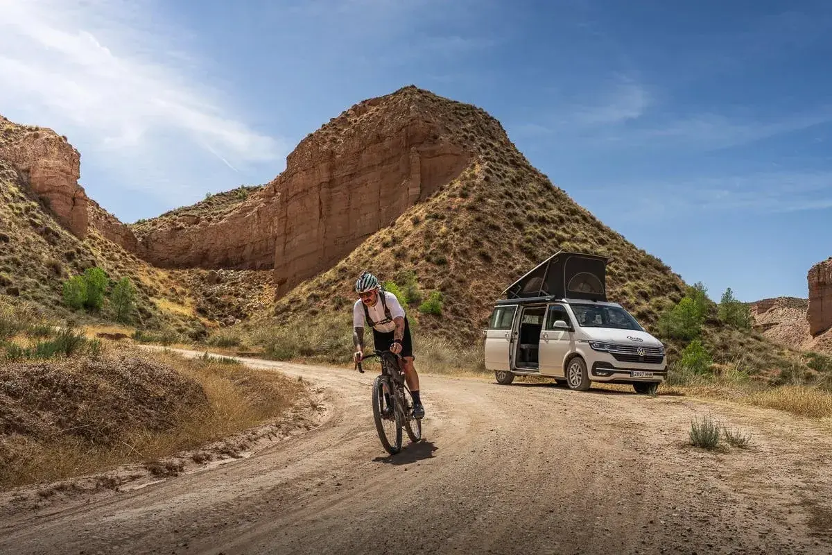 Ciclista pedaleando por un camino de badlands gravel, con una furgoneta camper aparcada al fondo.