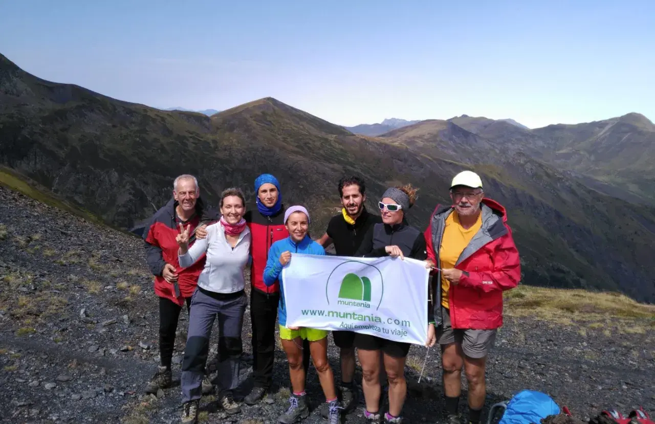 Grupo de senderistas celebra en la cima de una montaña, con el valle de **Aran** de fondo. Sostienen una pancarta de Muntania.com.