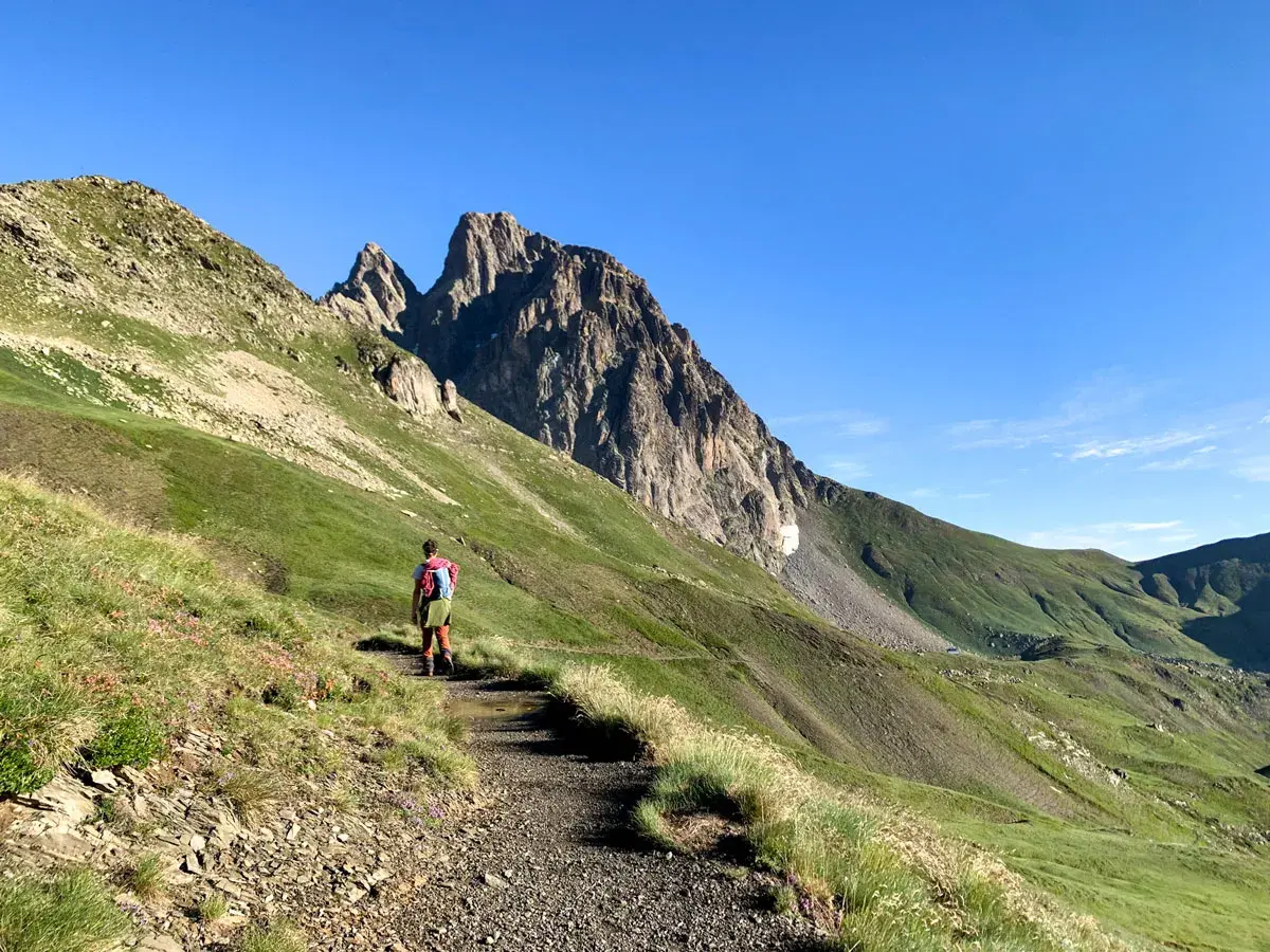 Senderista se dirige hacia el imponente Midi d'Ossau, rodeado de verdes laderas y un cielo azul despejado.