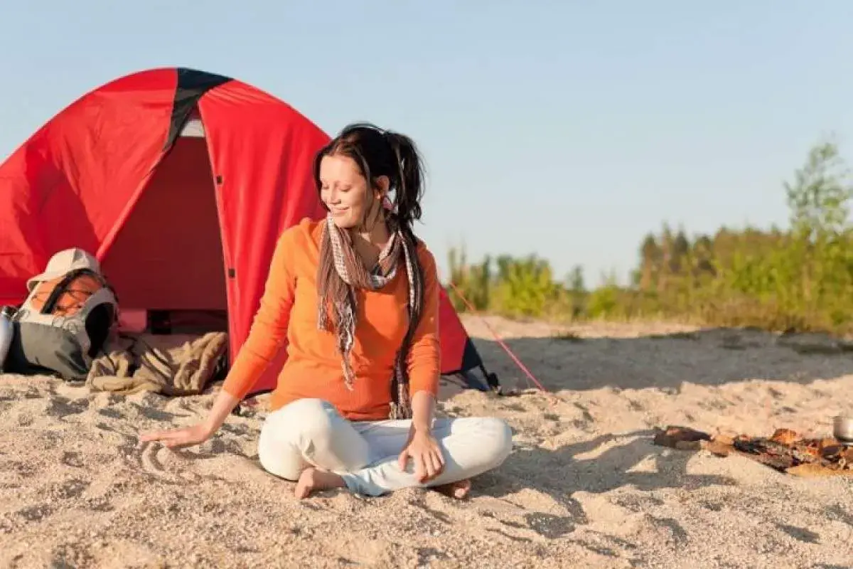 Mujer meditando junto a una tienda roja, disfrutando del acampar en la playa.