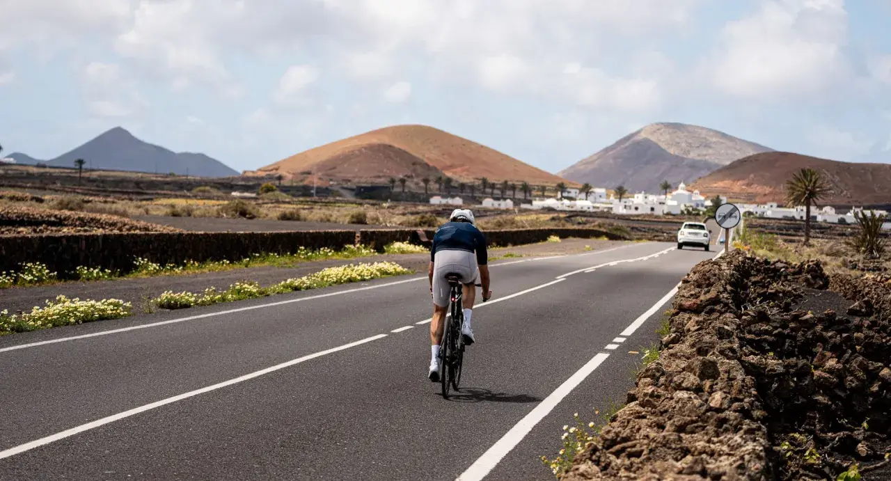 Ciclista pedaleando por una carretera en Lanzarote, con volcanes y casas blancas al fondo. Una aventura de lanzarote en bici.