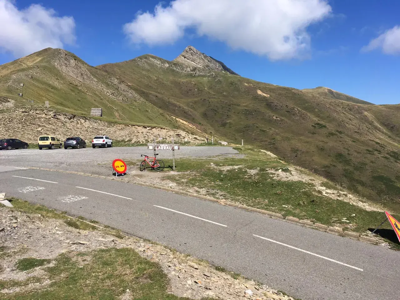 Un ciclista descansa junto a su bicicleta roja en el **puerto de Larrau**, con coches aparcados y montañas verdes bajo un cielo azul.
