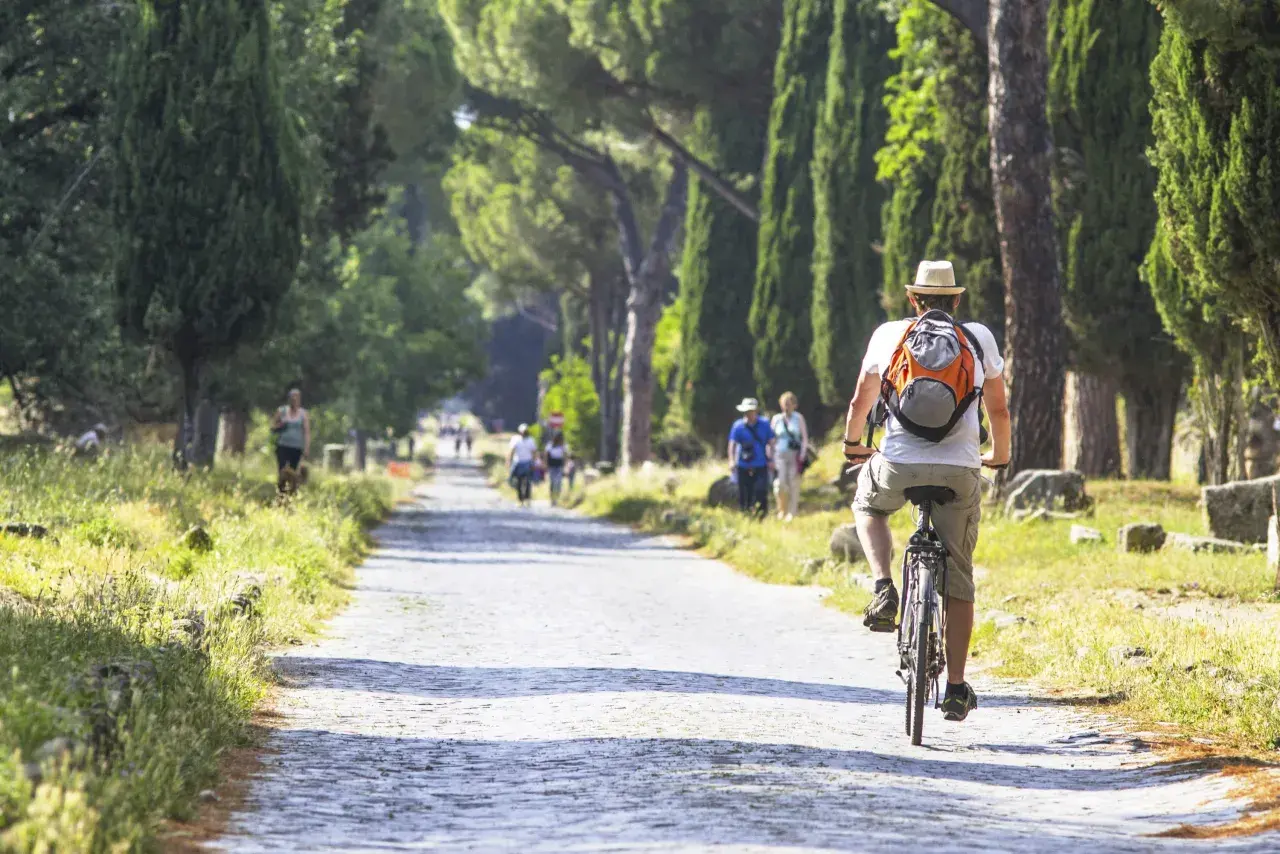 Un hombre pedalea por un camino empedrado en Roma, rodeado de altos cipreses y ruinas antiguas.
