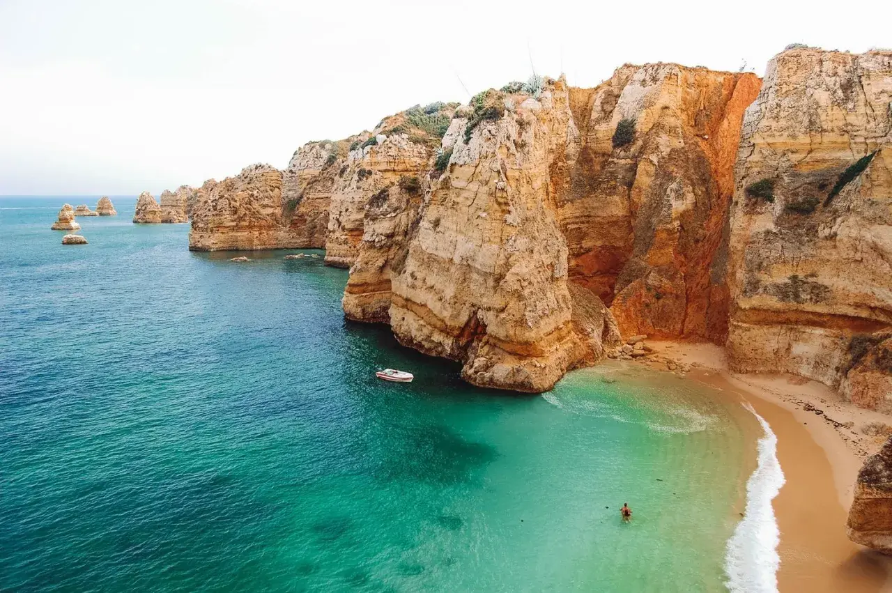 Aguas turquesas y acantilados dorados en las **playas Algarve**. Una pequeña barca navega cerca de una cala escondida donde una persona nada.