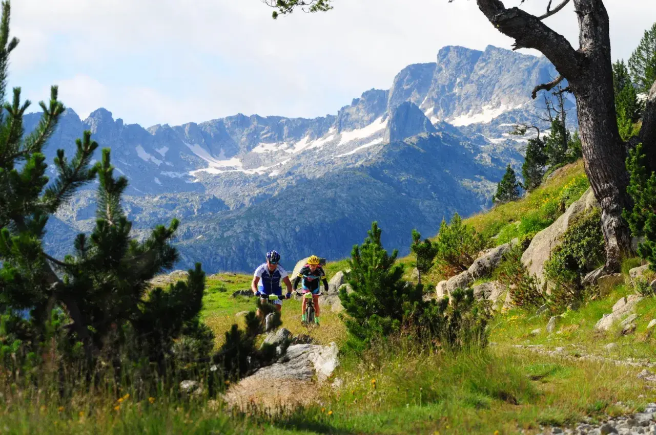 Ciclistas descienden por un sendero en el Pla de Beret, con imponentes montañas nevadas de fondo.
