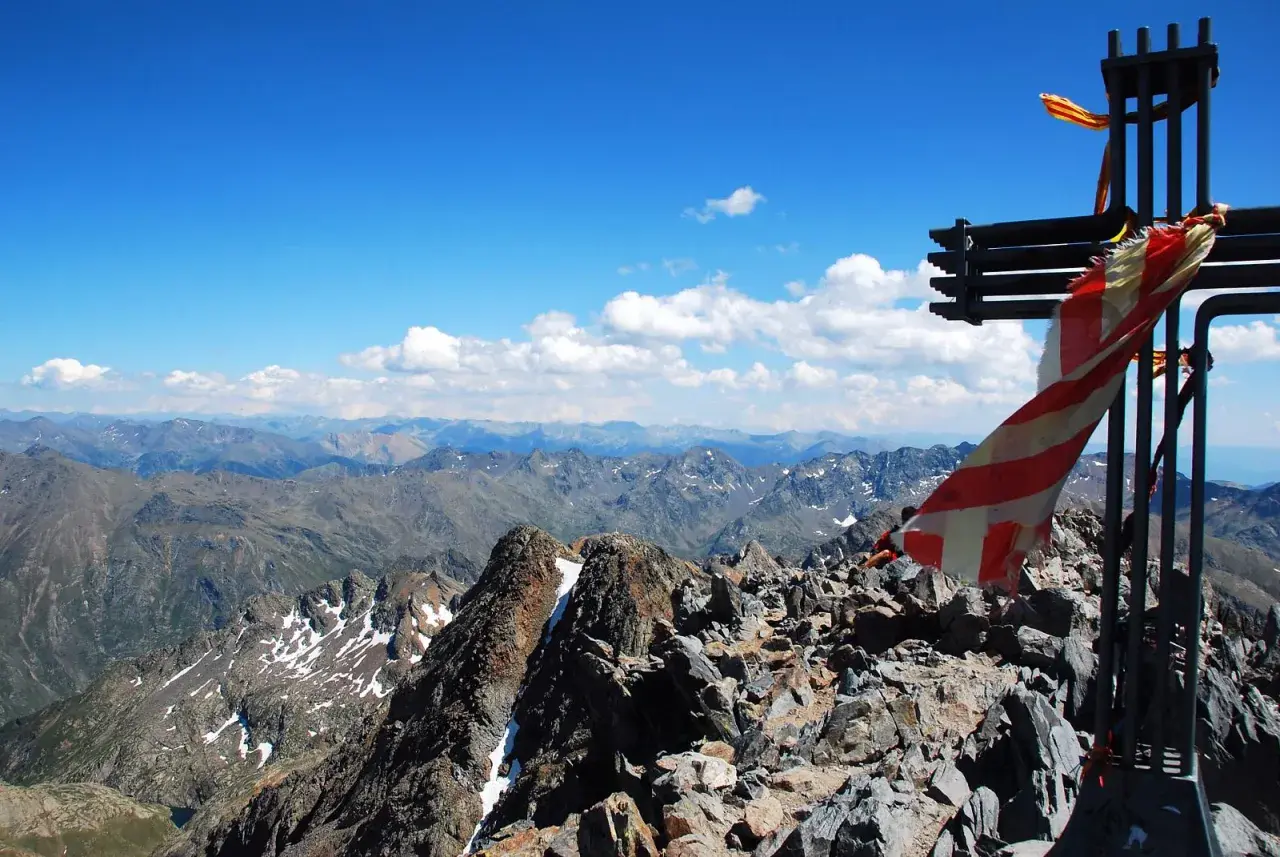 Cruz metálica en la cima de una pica d'estats, con bandera ondeando y un paisaje montañoso al fondo.