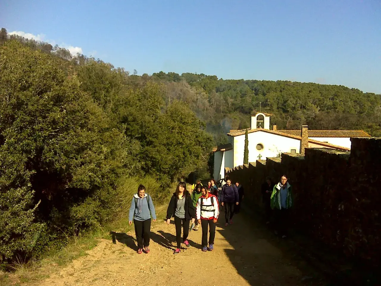 Un grupo de excursionistas camina por un sendero rural hacia una ermita blanca con un campanario. El paisaje es verde y montañoso, con árboles y vegetación. El sol brilla en un cielo azul claro. La ruta parece ser un camino de peregrinación, quizás hac...