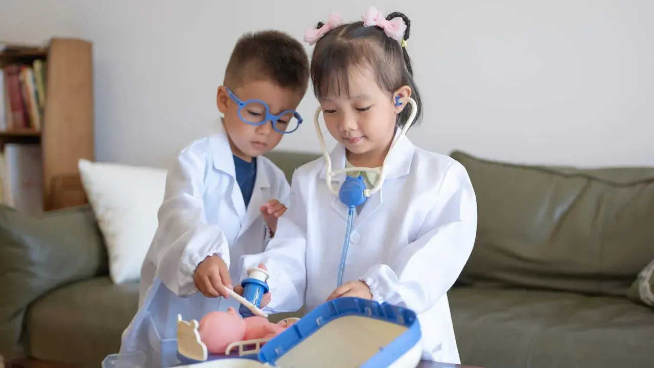 Dos niños, vestidos de doctores, atienden a un muñeco. La niña usa un estetoscopio, mientras el niño le administra una inyección. Es una escena de juego de roles, una brecha de Rolando con niños.