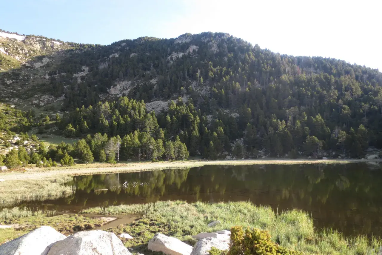 Paisaje sereno con un lago reflejando la montaña boscosa. El refugi de Malniu se esconde entre los árboles, invitando a la aventura.